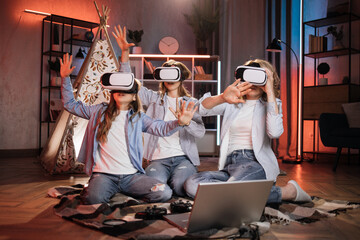 Happy mother and cute daughters sitting near toy wigwam at home and using modern VR headset during evening time. Caucasian family using modern technologies for playing at home. © sofiko14