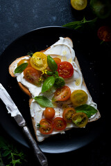 Cream cheese toast with cherry, colorful tomatoes. Black, moody background. Top view.