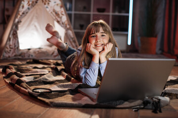Beautiful cute young girl on floor outside wigwam at home using laptop computer. Happy female spending evening time by watching favorite movie on digital device at home. © sofiko14