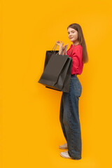 Beautiful young girl holds black paper bags after shopping on yellow background. Black Friday Concept. Vertical frame.