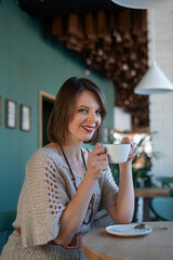 Young woman in gray dress drinks coffee on inside cafe. Portrait of brunette girl sits at table in cafe.