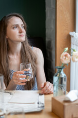 Young woman with glass of water sits at table in cafe and looks out the window waiting for an order. Portrait of girl in cafe.