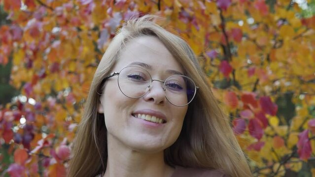 Woman enjoying autumn weather in the forest park. Cloudy day woods holiday journey. The face of a smiling happy girl with glasses holding red leaf. Rack focus shot.
