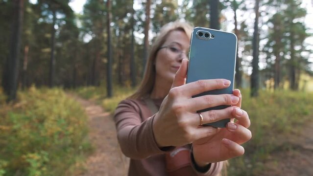 Blonde Girl blogger with glasses making selfie on a smartphone, outdoors in a forest park. Online, social lifestyle.
