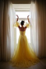 A woman's silhouette in a golden luxurious dress against the background of a window holds a curtain with her hands. Elegant lady in a yellow long silk dress with bare back, back view.