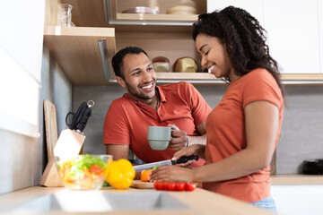 Cheerful young black couple in same t-shirts talking, wife preparing salad, man drink coffee