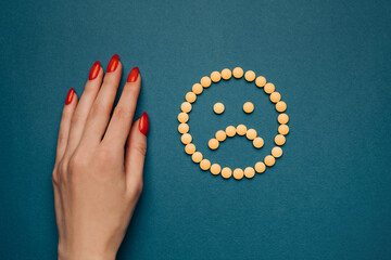 Obraz premium A woman's hand with a red manicure and pills are laid out on a blue background in the form of a sad emoticon. Top view. Antidepressants concept or headache.