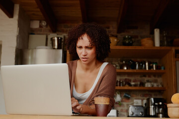 Young biracial woman frowning while working on laptop in kitchen at home