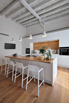 interior view of a modern white kitchen, in the foreground the island kitchen with stools, the lighting is entrusted to four pendant lamps, the floor and ceiling are made of wood