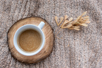 Cup of coffee cappuccino on tree cut wooden cup coaster, beige sweater and dry autumn leaves on table background. Cozy beige autumn, fall aesthetic. Top view, flat lay, copy space