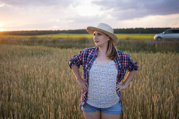 Obraz premium A beautiful middle-aged farmer woman in a straw hat and a plaid shirt stands in a field of golden ripening wheat during the daytime in the sunlight