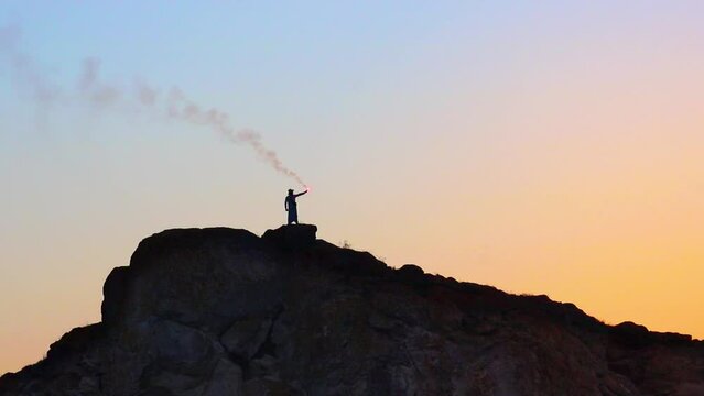 An unrecognizable man on a mountain holds a smoking signal flare in his hand, against the backdrop of a sunset. Signal checker, smoke chip, fireworks and fire.