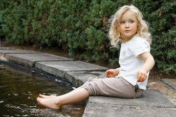 Portrait of blue-eyed little girl with long curly fluffy hair sitting on concrete edge of pond with...
