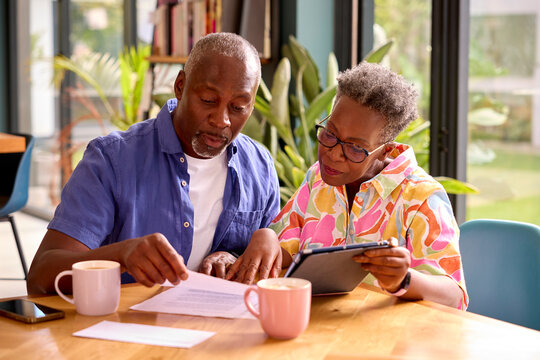 Senior Couple Sitting Around Table At Home Reviewing Finances Using Digital Tablet