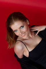 Vertical smiling blonde woman in black dress and capote look camera and posing in red studio. Mascara, eyeliner, shades