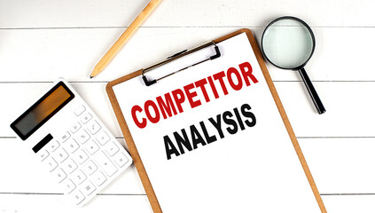 COMPETITOR ANALYSIS words on clipboard, with calculator, magnifier and pencil on the white wooden...