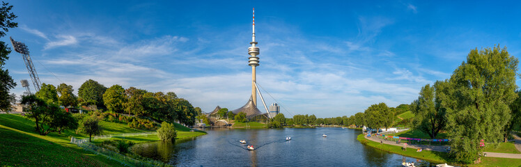 Panoramafoto vom Olympiapark in München mit Olympiaturm, See, Laubbäumen und blauem Himmel bei...