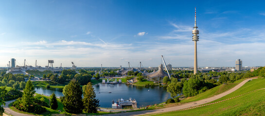 Panoramafoto vom Olympiapark in München mit Olympiaturm, See, Laubbäumen und blauem Himmel bei sonnigem Sommerwetter