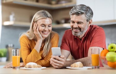 Happy beautiful middle aged spouses having breakfast, checking smartphone