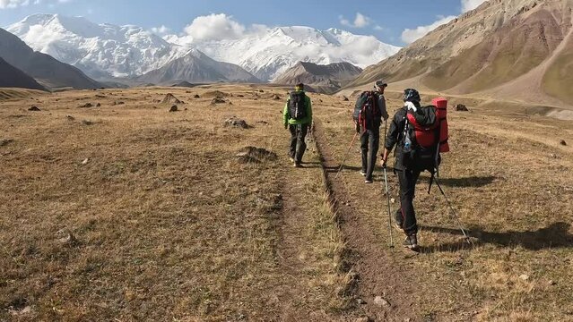 Three tourists with backpacks are walking along the plain along the mountains. Trekking from Base Camp to Camp 1 under Lenin Peak, Kyrgyzstan. Beautiful summer mountain landscape.
