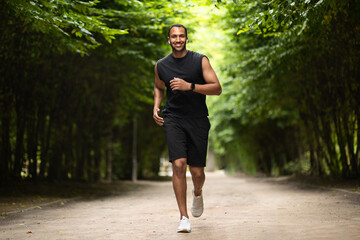 Motivated african american athletic guy jogging by green public park