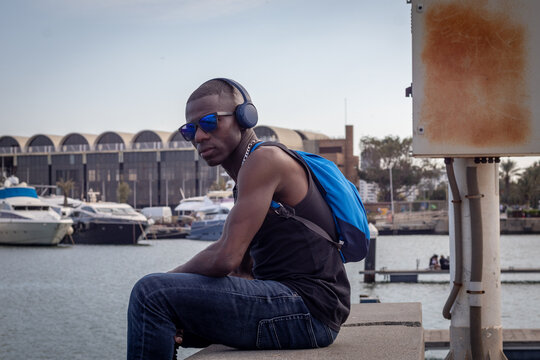 Hombre africano con gafas de sol azules y morral azul, en el puerto escuchando musica. 