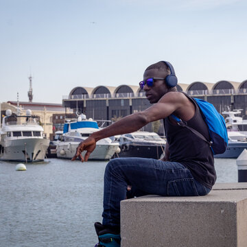 Hombre africano con gafas de sol azules y morral azul, en el puerto escuchando musica. 