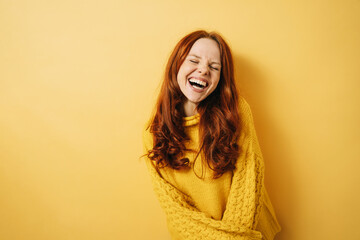 Laughing redhead woman with closed eyes and yellow sweater, copyspace against yellow background - obrazy, fototapety, plakaty
