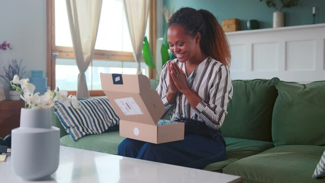Young enthusiastic casual African American woman unpacks gift from friends by opening cardboard box and feels joy or does unboxing after shopping in online store sits on couch at home
