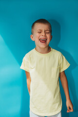 A 6-7-year-old boy in a yellow T-shirt on a blue background shouts loudly. Portrait of a child, front view