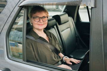 Business woman in glasses is sitting in the back seat of a car. Business woman is working on laptop in car. Middle aged woman with glasses smiles and looks into the camera.