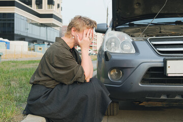 Middle-aged woman sits on the side of road in front of broken-down car with an open hood. Business lady is sad about the breakdown of her car. Female driver needs technical assistance on the road.