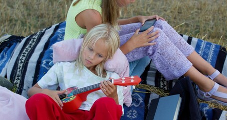 Fototapeta premium charming girl playing the ukulele on mown rye in field. Landscape of straw bales against setting sun on background. Happy sisters watch video on smartphone outdoors. Childhood. Country life
