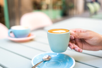 woman's hand holding a cup of coffee with milk. she is recharging her energy by drinking coffee on the terrace of the store. the cup and saucer are colorful. photo with copy space.