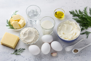 Ingredients for cooking cheese gougères: eggs, milk, water, butter, flour, hard cheese, olive oil, salt, black pepper, cream cheese, fresh dill, garlic on a light blue background