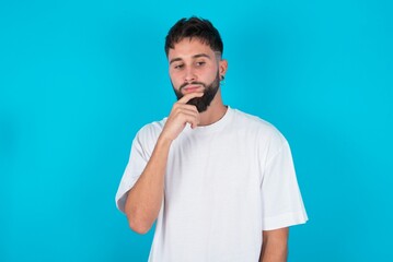 Shot of contemplative thoughtful bearded caucasian man wearing white T-shirt over blue background keeps hand under chin, looks thoughtfully upwards.