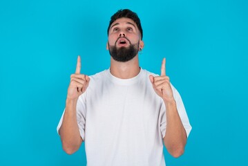 bearded caucasian man wearing white T-shirt over blue background being amazed and surprised looking and pointing up with fingers showing something strange.