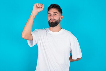 bearded caucasian man wearing white T-shirt over blue background feeling serious, strong and rebellious, raising fist up, protesting or fighting for revolution.