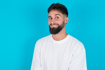 bearded caucasian man wearing white T-shirt over blue background with thoughtful expression, looks away keeps hands down bitting his lip thinks about something pleasant.