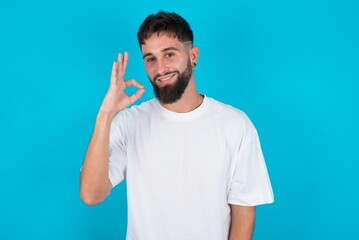 Glad attractive bearded caucasian man wearing white T-shirt over blue background shows ok sign with hand as expresses approval, has cheerful expression, being optimistic.