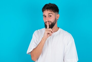 bearded caucasian man wearing white T-shirt over blue background makes hush gesture, asks be quiet. Don't tell my secret or not speak too loud, please!