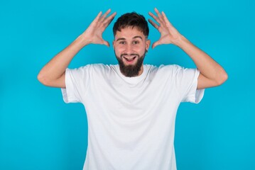 Fototapeta na wymiar bearded caucasian man wearing white T-shirt over blue background looks with excitement at camera, keeps hands raised over head, notices something unexpected, reacts on sudden news.