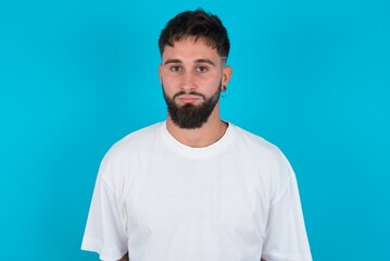 Fototapeta premium Joyful bearded caucasian man wearing white T-shirt over blue background looking to the camera, thinking about something. Both arms down, neutral facial expression.