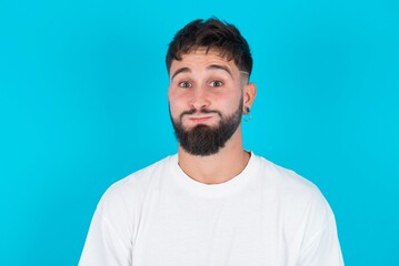 bearded caucasian man wearing white T-shirt over blue background puffing cheeks with funny face. Mouth inflated with air, crazy expression. - obrazy, fototapety, plakaty