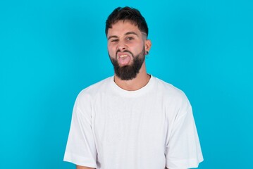 Body language. Disgusted stressed out bearded caucasian man wearing white T-shirt over blue background, frowning face, demonstrating aversion to something.