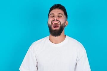 Stressful bearded caucasian man wearing white T-shirt over blue background screams in panic, closes eyes in terror, keeps hands on head, finds out terrified news, can't believe it.