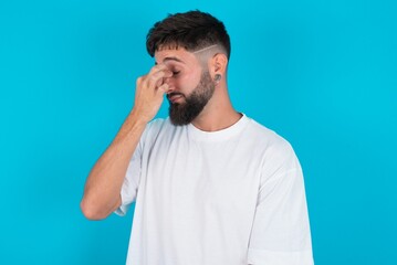 Very upset, bearded caucasian man wearing white T-shirt over blue background touching nose between closed eyes, wants to cry, having stressful relationship or having troubles with work