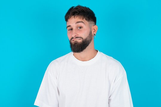 bearded caucasian man wearing white T-shirt over blue background being nervous and scared biting lips looking camera with impatient expression, pensive.