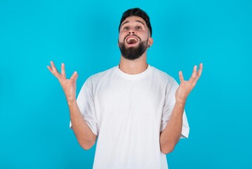 Joyful excited lucky bearded caucasian man wearing white T-shirt over blue background cheering, celebrating success, screaming yes with clenched fists