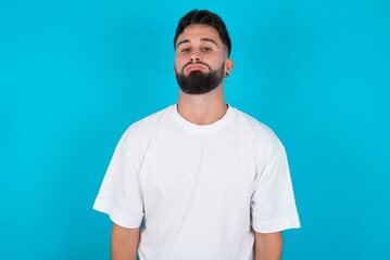 Gloomy, bored bearded caucasian man wearing white T-shirt over blue background frowns face looking up, being upset with so much talking hands down, feels tired and wants to leave.